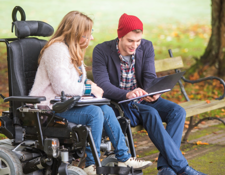 A blonde woman in a wheelchair chats to a man with a red woolly hat who is sitting on a bench and using a tablet