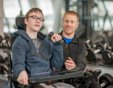Young man in wheelchair, holding an exercise weight, with a trainer